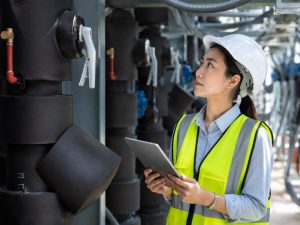 Ingeniera con casco y chaleco de seguridad revisa una tablet frente a equipos y tuberías en una instalación industrial