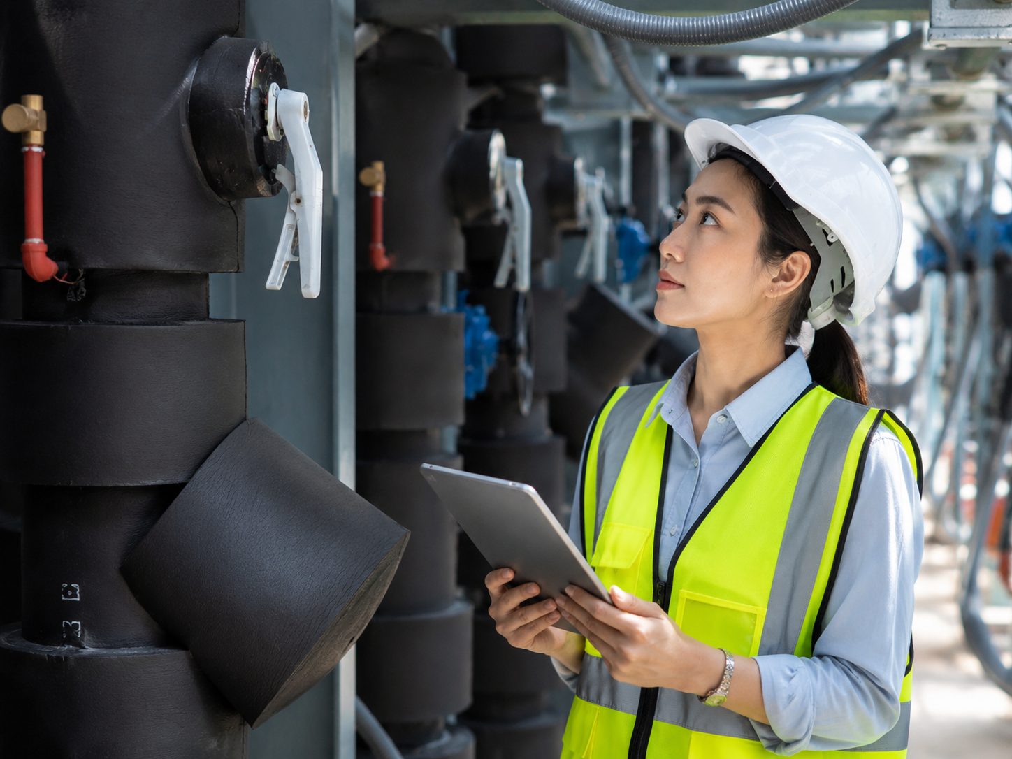 Ingeniera con casco y chaleco de seguridad revisa una tablet frente a equipos y tuberías en una instalación industrial