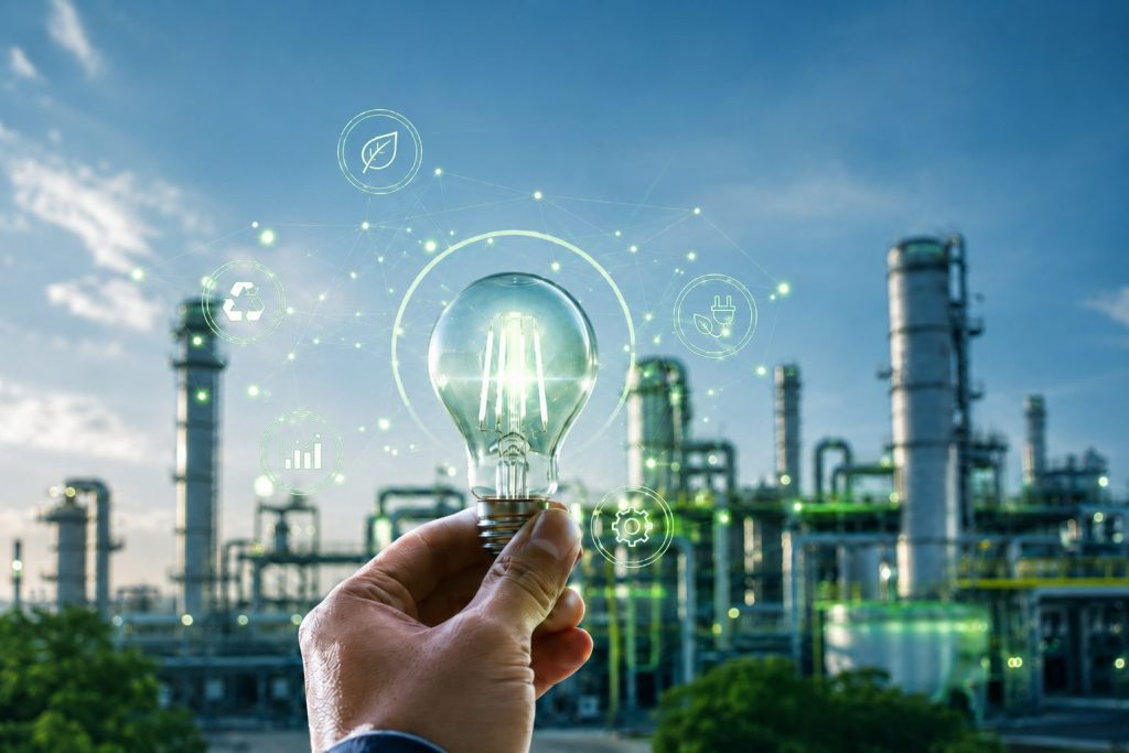 Imagen conceptual de eficiencia energética industrial, con una bombilla iluminada frente a una planta industrial y elementos digitales vinculados a sostenibilidad, ahorro energético, optimización de procesos y descarbonización.