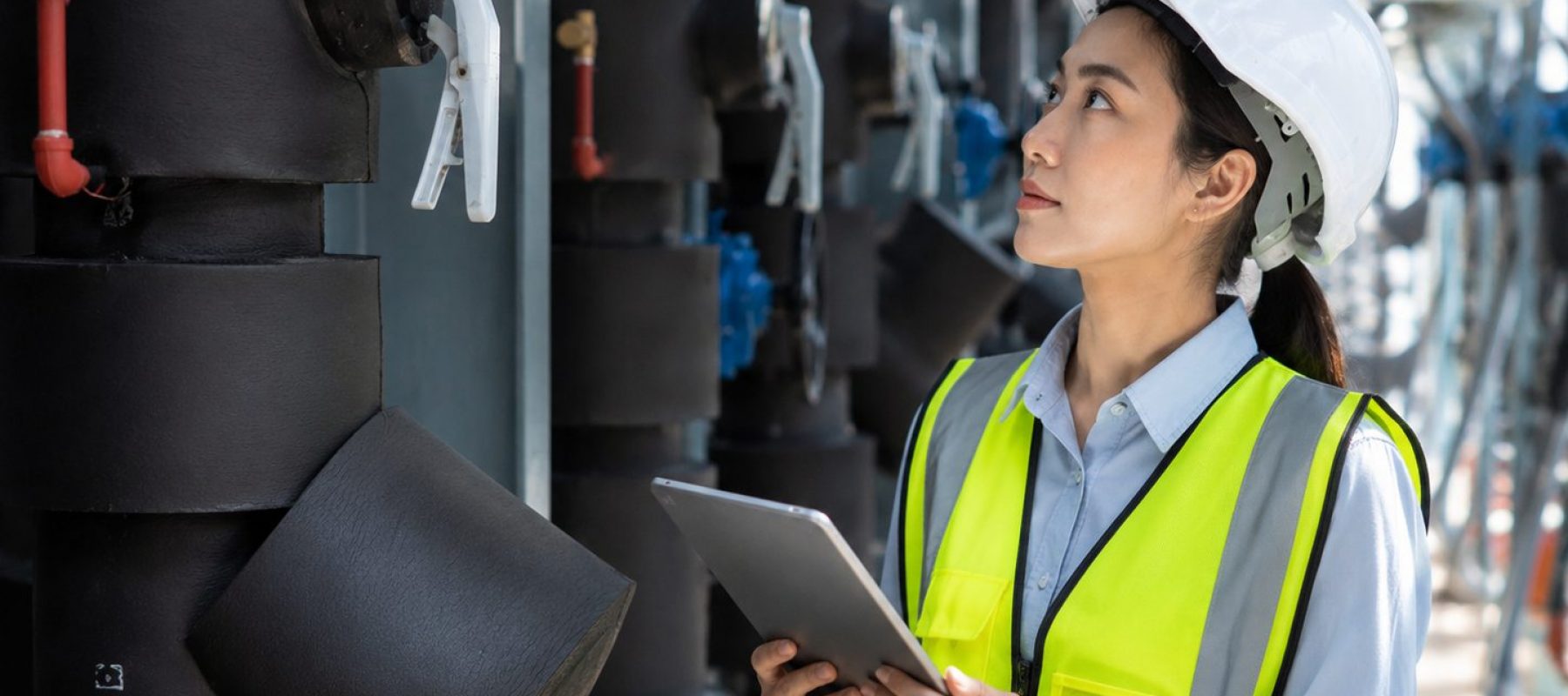 Ingeniera con casco y chaleco de seguridad revisa una tablet frente a equipos y tuberías en una instalación industrial